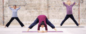 The image shows three individuals performing Angamardana yoga exercises in an outdoor setting against a stone wall. The person in the center is bent forward with legs spread wide apart, hands resting on the ground, and head down. The two individuals on either side are standing with legs spread wide and hands clasped behind their heads. They are all on yoga mats and appear to be engaged in a synchronized yoga session. This image is interesting and relevant as it depicts a group practicing Angamardana, a traditional Indian fitness system that involves a series of physical postures and movements aimed at strengthening the body and improving flexibility.
