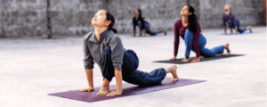 A group of individuals practicing yoga in an outdoor setting, each in a Ashvasanchalana pose, focusing on balance and flexibility. The participants, dressed in comfortable yoga attire, are on mats placed on a concrete floor with a simple, gray stone wall in the background. The image conveys focus, strength, and flexibility achieved by practicing Surya kriya & Surya shakti yogic practices.