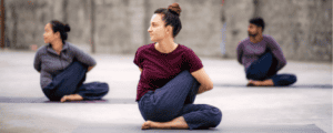 The image shows three individuals practicing yogasanas. They are seated on yoga mats in a cross-legged position with their torsos twisted to one side and their arms wrapped around their backs, performing a seated spinal twist pose. The background is a plain, outdoor setting with a concrete floor and a blurred wall. The individuals are dressed in comfortable, casual clothing suitable for yoga practice. This image is interesting and relevant as it depicts a common yoga pose that promotes flexibility and spinal health.