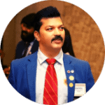 The image shows an individual wearing a blue suit jacket, a white shirt, and a red tie with diagonal stripes. The individual has several pins and a name tag on the left side of the suit jacket. The background appears to be an indoor setting with another person partially visible behind the main subject. This image is used for a positive testimonial, likely highlighting the individual's professional appearance and achievements.
