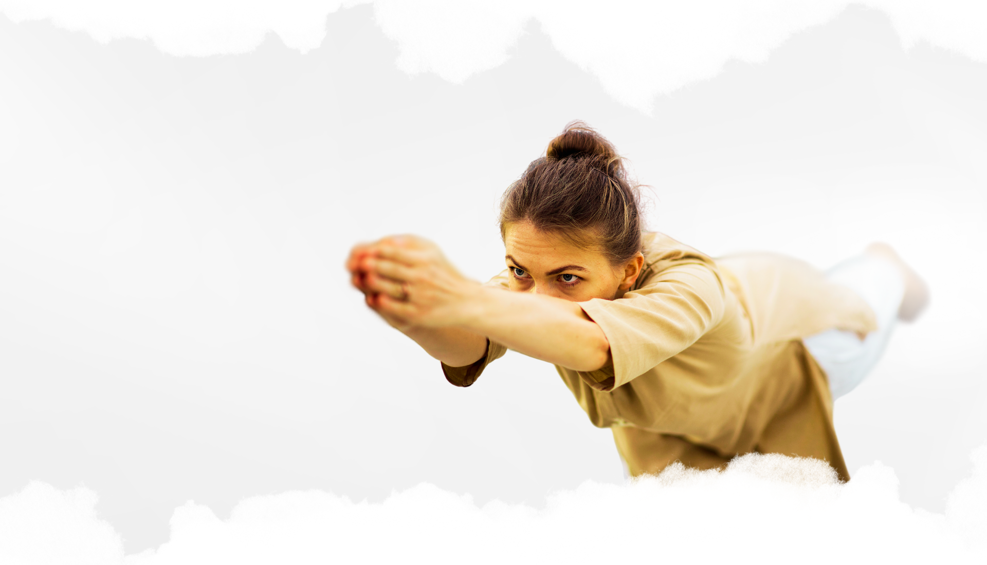 This image shows a focused woman in mid-pose, practicing a dynamic yoga posture that emphasizes strength, balance, and concentration. She leans forward with arms extended in front of her, gazing intensely ahead. Her body is engaged, symbolizing physical and mental discipline, and she is dressed in loose, neutral-toned clothing that allows ease of movement. The background is a soft, light gradient with a white, cloud-like border around the edges, creating a sense of calm and spaciousness. This image likely represents the practice of Angamardana, a traditional yogic sequence aimed at enhancing physical fitness and inner focus.